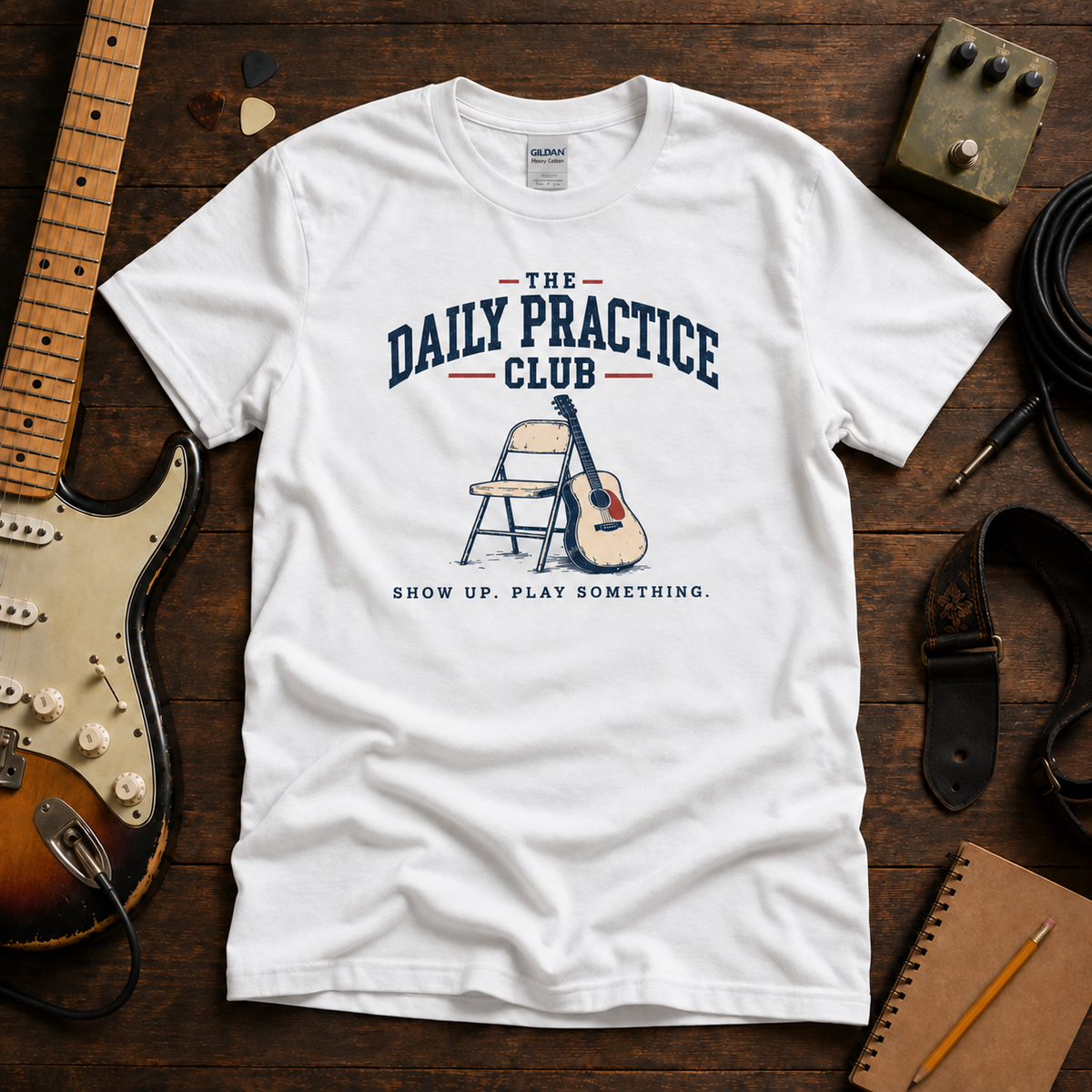 White Daily Practice Club tee by Steady Strum, laid flat on a wood desk with guitar gear