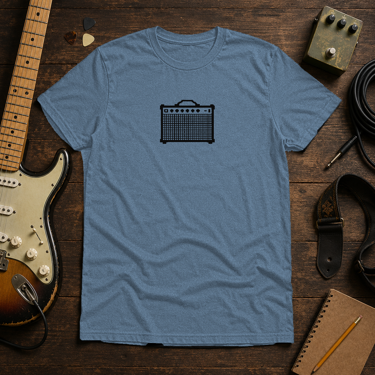 Heather Indigo Pixel Amp tee by Steady Strum, laid flat on a wood desk with guitar gear