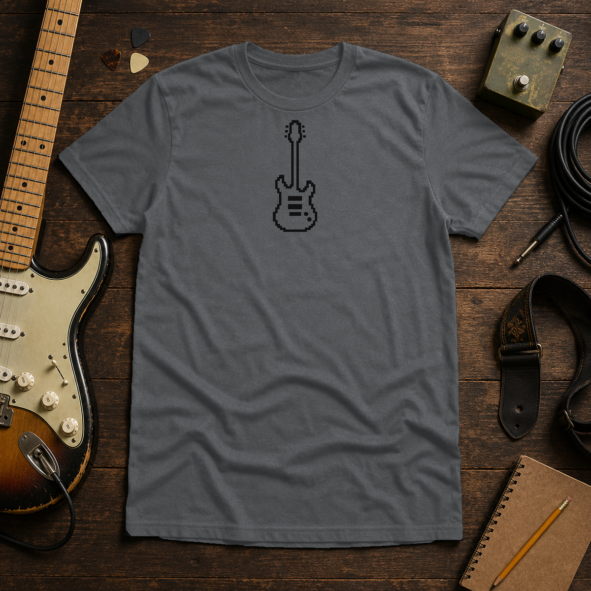 Graphite Heather Pixel Guitar tee by Steady Strum, laid flat on a wood desk with guitar gear