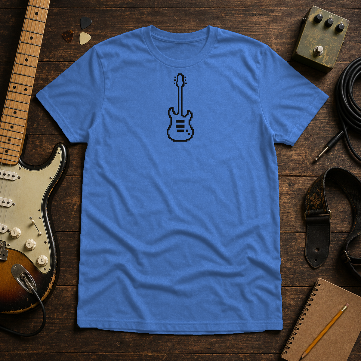 Heather Royal Pixel Guitar tee by Steady Strum, laid flat on a wood desk with guitar gear