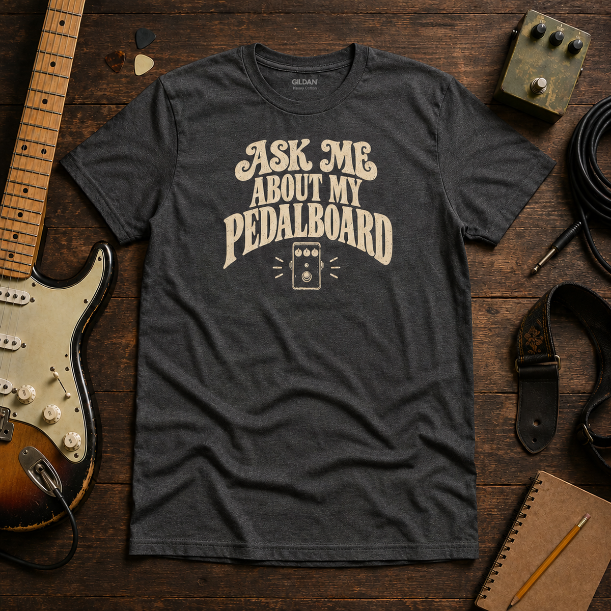 Dark Grey Ask Me About My Pedalboard tee by Steady Strum, laid flat on a wood desk with guitar gear