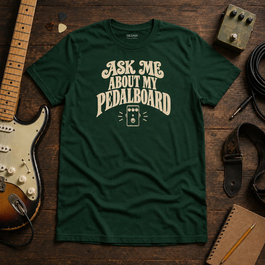 Green Ask Me About My Pedalboard tee by Steady Strum, laid flat on a wood desk with guitar gear