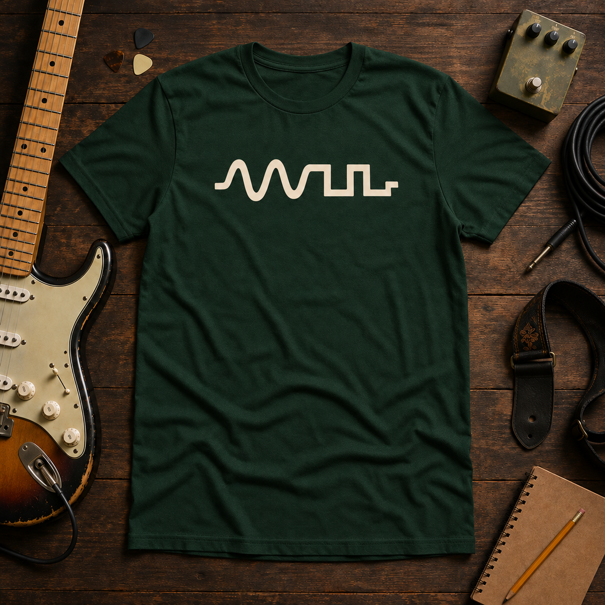 Green Clipped Wave tee by Steady Strum, laid flat on a wood desk with guitar gear