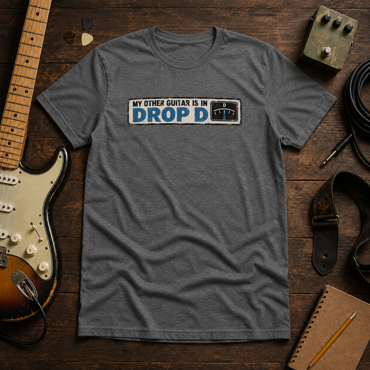 Graphite Other Guitar Is In Drop D tee by Steady Strum, laid flat on a wood desk with guitar gear