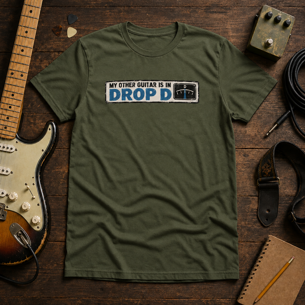 Green Other Guitar Is In Drop D tee by Steady Strum, laid flat on a wood desk with guitar gear