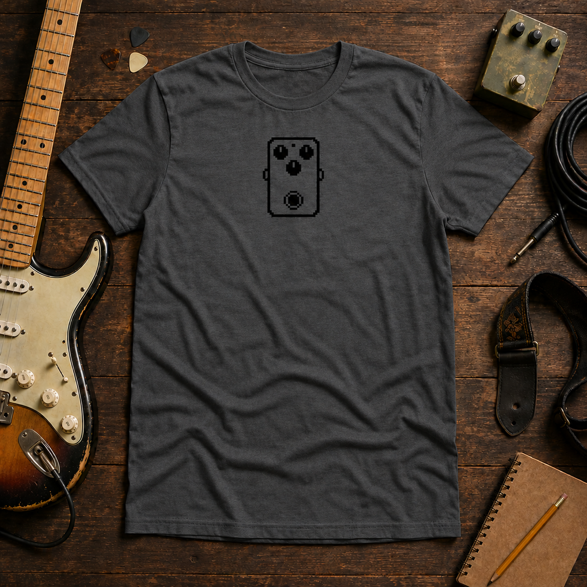 Charcoal Pixel Pedal tee by Steady Strum, laid flat on a wood desk with guitar gear