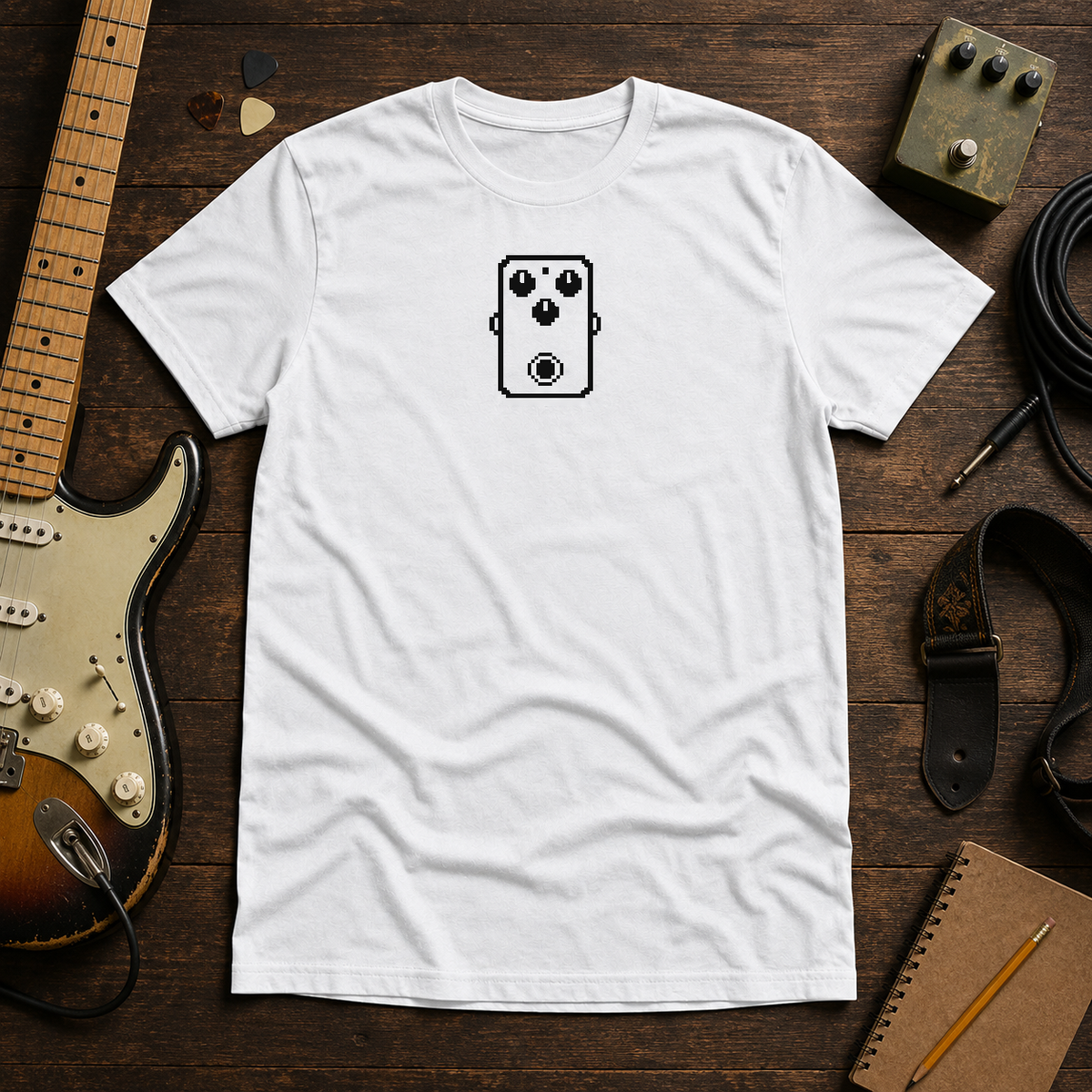 White Pixel Pedal tee by Steady Strum, laid flat on a wood desk with guitar gear